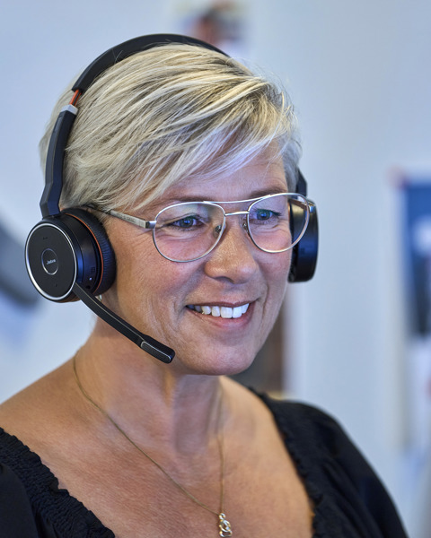 A smiling woman with short blonde hair and glasses, wearing a black headset with a microphone