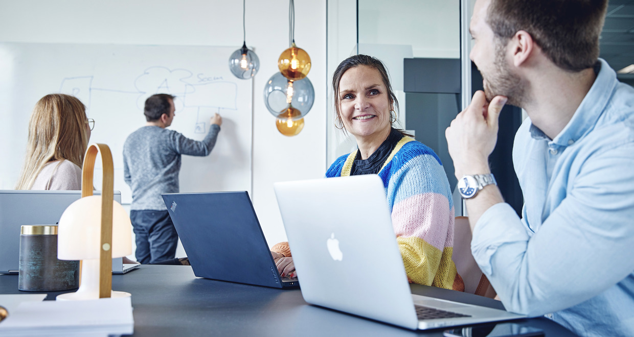 Smiling employees at Danoffice IT collaborate in a modern office, in a meeting room, with one person writing on a whiteboard and others working on laptops